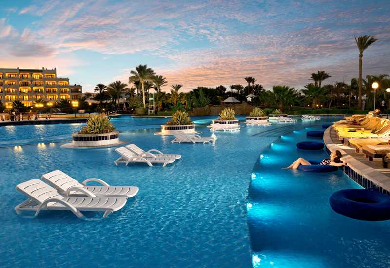 Verlicht zwembad met luxe ligbedden in het water tijdens een sfeervolle zonsondergang op vakantie.